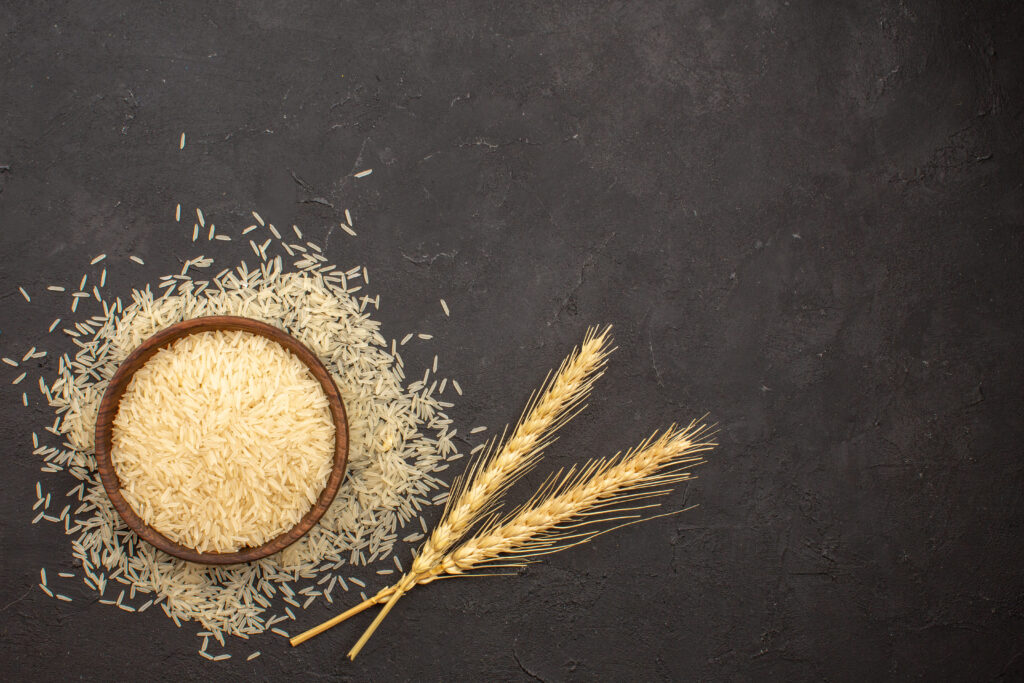 A top-down view of raw rice in a wooden bowl, surrounded by scattered grains and two wheat stalks, on a dark surface.